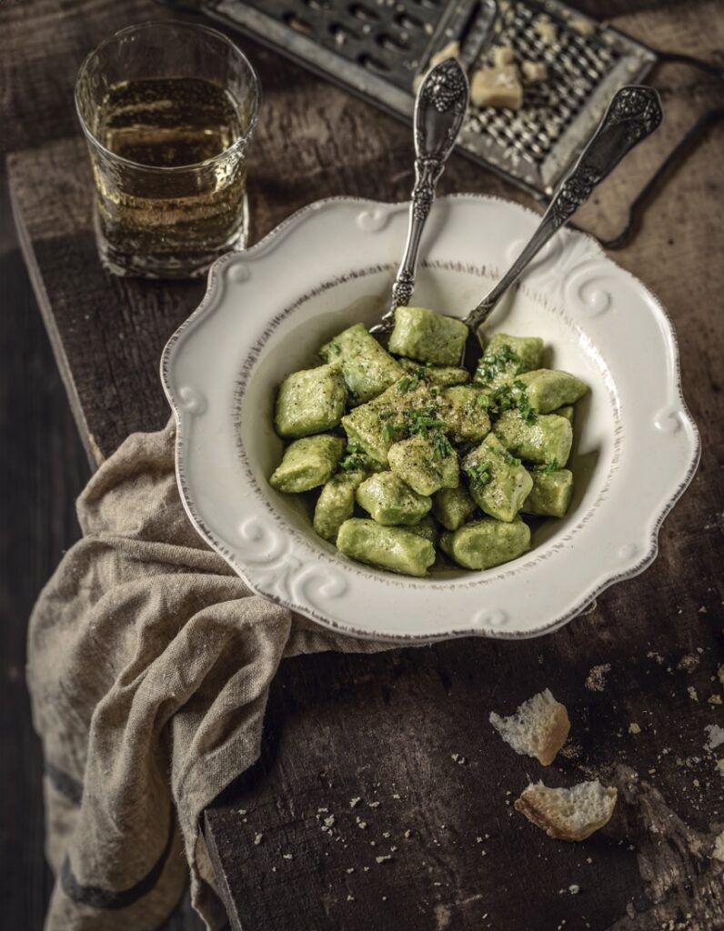 A white bowl of Potato and Pea Gnocchi with two forks in the bowl sits on a wood table.