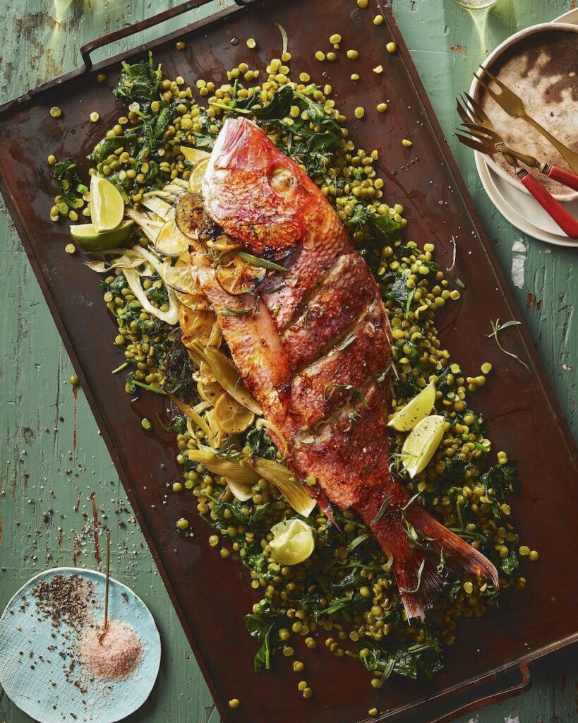 A large Red Snapper lays on a cooking sheet surrounded by a Green Spit Pea Salad and limes in celebration of Nowruz.