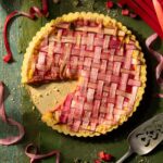 A Rhubarb Frangipane Tart with a slice taken out of it featuring latticed rhubarb on top and around the tart on the green table.