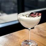 A cocktail glass holds a white Snow White's Wicked Apple Cocktail with a white foam on top and a dehydrated apple and gold flake garnish.