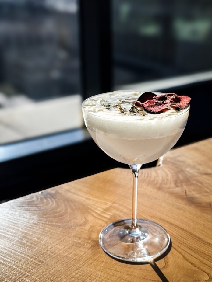 A cocktail glass holds a white Snow White's Wicked Apple Cocktail with a white foam on top and a dehydrated apple and gold flake garnish.