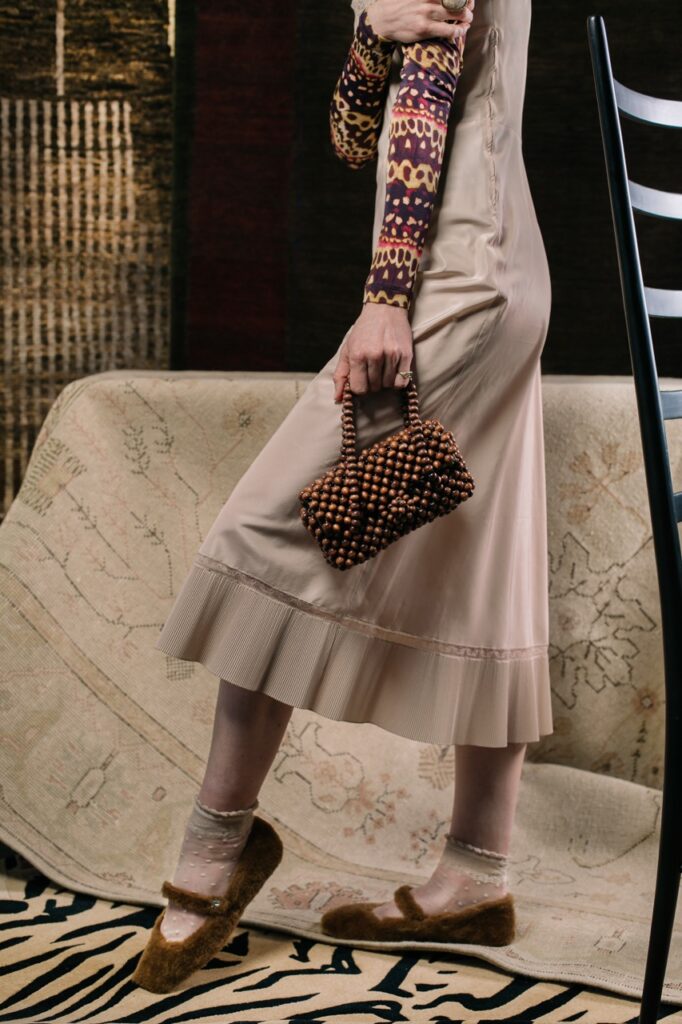A woman in a long neutral dress holds a brown clutch bag in her hand with brown sandals on to match.