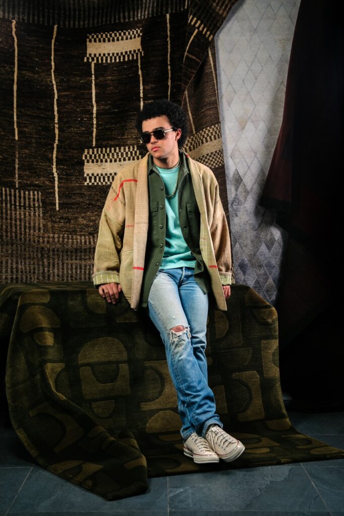 A man leans against a table in layered spring jackets and jeans with sunglasses on as well.