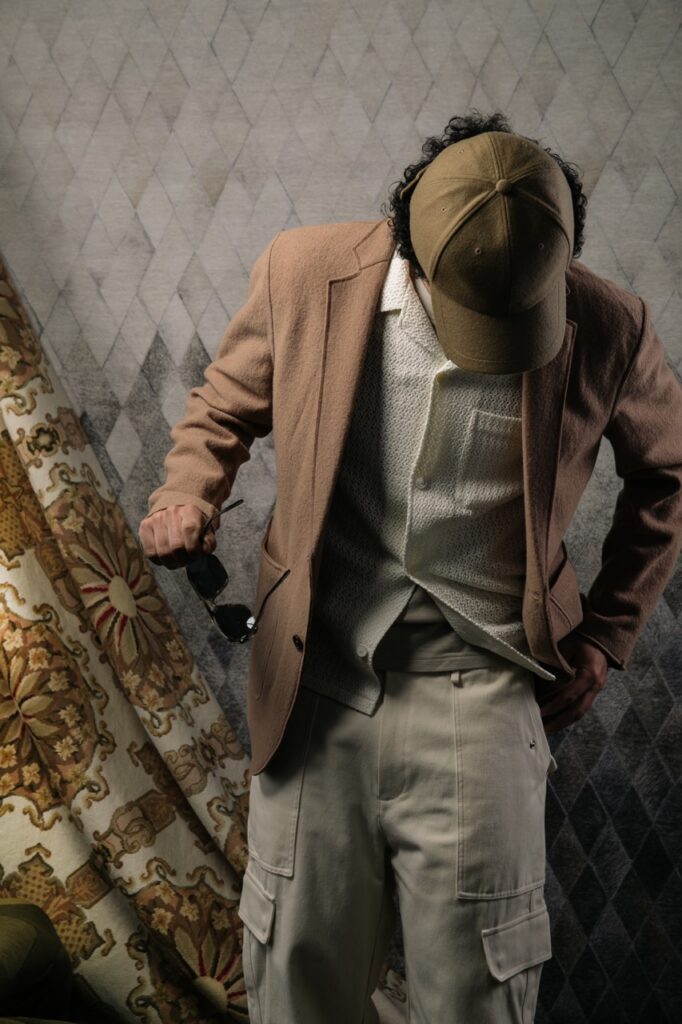 A man looks down wearing a brown hat, brown blazer and holding brown sunglasses in his hand.
