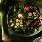 A big teal bowl of springtime vegetable succotash using radishes and various green sprouts as a green onion sits to the left of the bowl.