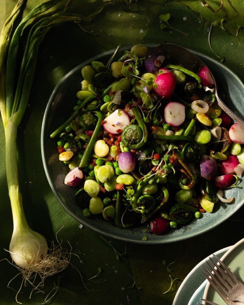 A big teal bowl of springtime vegetable succotash using radishes and various green sprouts as a green onion sits to the left of the bowl.
