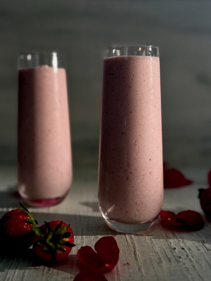 Two strawberry boozy smoothies sit on a white background, slightly pink in color with rose petals and strawberries around the base of the glasses.