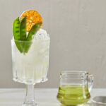 A sweet pea cocktail in a glass garnished with pea pods and an orange whel.