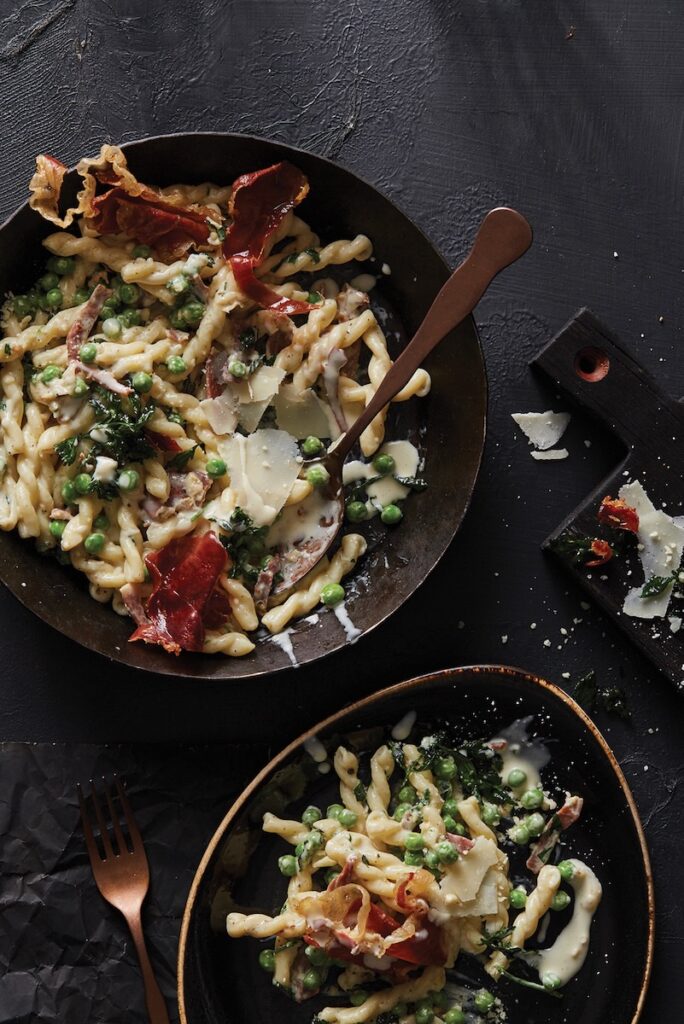 Gemelli, prosciutto and peas in a saucepan on a black background.
