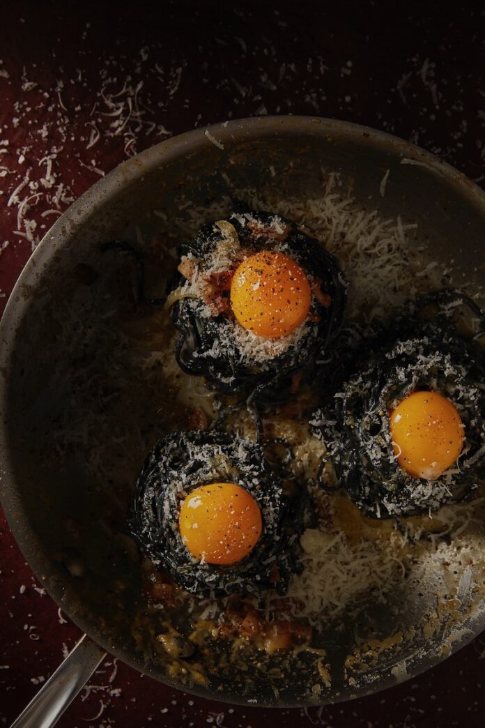 Three bundles of handmade charcoal trenette made intoa. traditional Carbonara with yellow cheese on top of each pile.