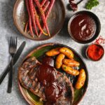A large plate holds a steak, potatoes, and veggies with a red chile sauce over top of it all on a white table.