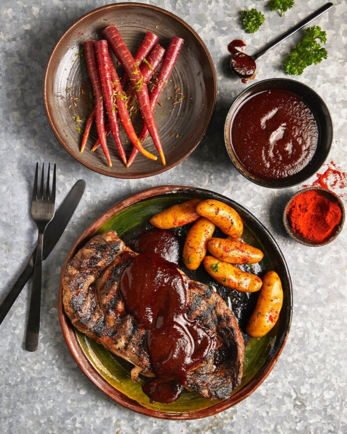 A large plate holds a steak, potatoes, and veggies with a red chile sauce over top of it all on a white table.