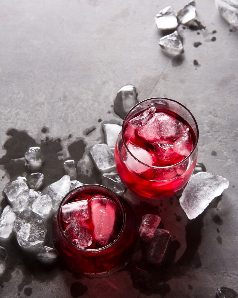 Two glasses of chilled red wine with ice cubes in them sit on a grey table with ice cubes scattered about.