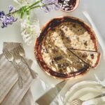 A Chocolate Chip Cake with chocolate ganache on top sits on a white table with white and purple flowers laying near by and a silver serving spatula.