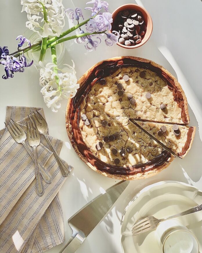 A Chocolate Chip Cake with chocolate ganache on top sits on a white table with white and purple flowers laying near by and a silver serving spatula.