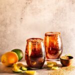 Two Dark and Handsome Rum Moscow Mules sit in front of a painted brown background with Tajin rims and a lime and orange sitting next to the glasses.
