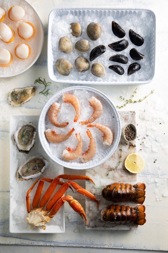 Various shellfish sit in groups on ice and platters such as raw shrimp, snow crab claws, oysters, scallops, lobster tails, and mussels.