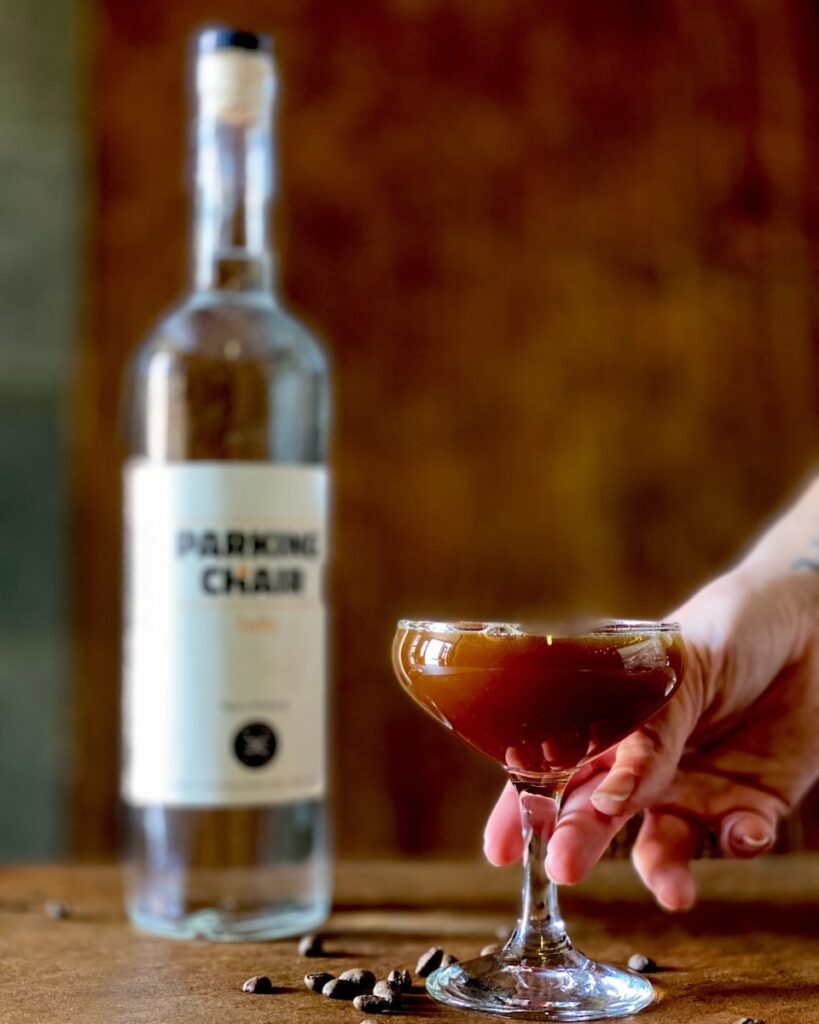 A person places down a coupe glass with an espresso martini in it as a bottle of parking chair vodka sits blurred in the background.