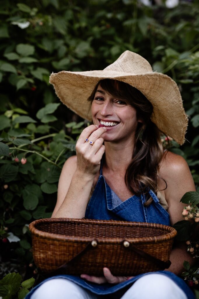 A woman in a sunhat sits and eats backyard raspberries from a basket on her lap.
