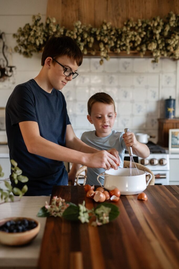 A young boy and his older brother mix something a large pot on a wood kitchen counter.