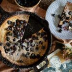 A dutch baby with blueberries sits in a cast iron pan with a slice on a plate next to it.