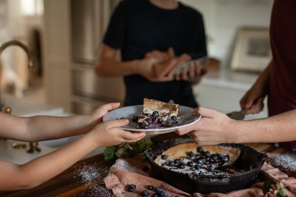 A person hands another a plate of a slice of blueberries on top of a dutch baby.