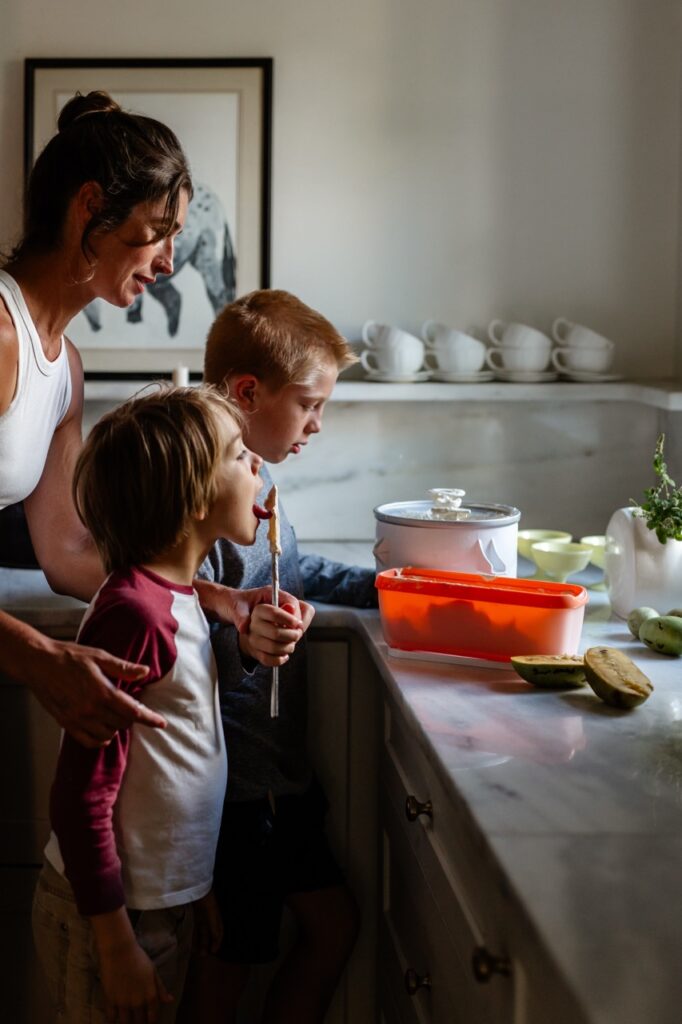 A young boy licks a spoon while standing with his mother and brother in the kitchen.