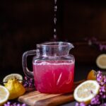 A glass pitchers of redbud lemonade with halved lemons surrounding the pitcher.