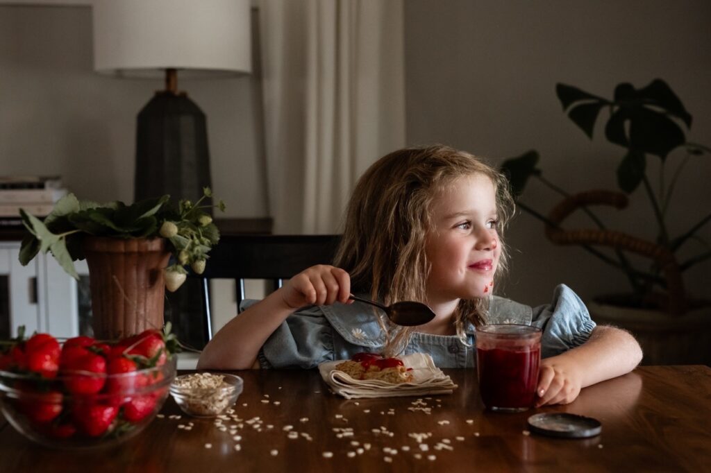 A little girl holds a spoon of strawberry jam as oats sit scattered on the table in from of her.
