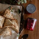 Oat scones and jars of strawberry jam from garden strawberries sit on a wood table.