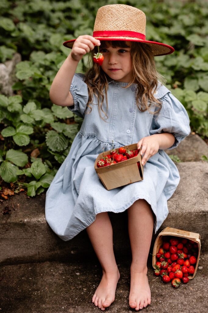 A young girl in a blue dress and red sunhat sits on a bench holding a garden strawberry in front of her face with a basket of strawberries on her lap.