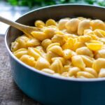 A blue pot of cheddar shell mac and cheese in a yellow color with a wooden spoon stuck inside it.