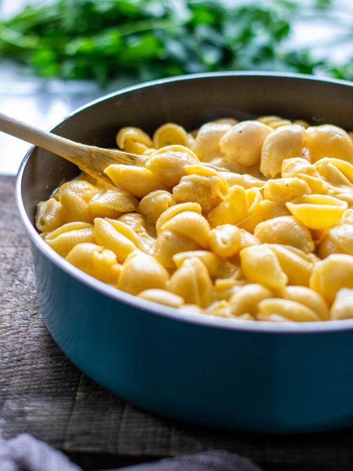 A blue pot of cheddar shell mac and cheese in a yellow color with a wooden spoon stuck inside it.