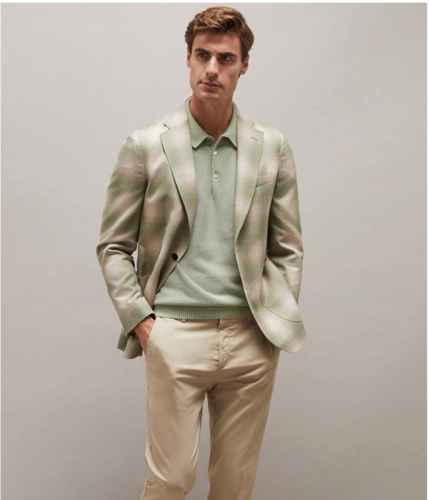 A man in a cream and soft green three piece suit stands against a neutral background with his hands in his pocket.
