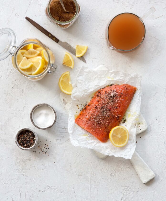 A piece of salmon to be grilled sits on white butcher paper surrounded by lemon slices, a citrus shrub in a glass, and a small container of sugar.