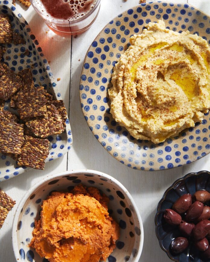 Two bowls of hummus, one lemon artichoke and the other triple tomato sit on a white picnic table with wheat crisps nearby.