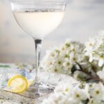 In a coupe glass on a table cloth sits a Lavender Lemon Drop for Mother's Day as white flowers and a lemon twist lay beside the glass.