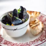 A small bowl hols Mussels in a broth that sit on a plate with two pieces of toasted baguette beside the bowl.