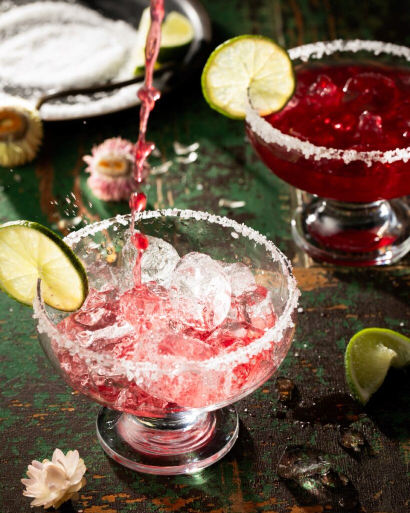 A pomegranate margarita gets poured into a margarita glass full of ice with salt and a lime wheel on the rim as a completed cocktail sits behind it on a green table.
