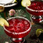 Two margarita glasses with salt rims are filled with a red pomegranate margarita with a lime wheel garnishing each glass.