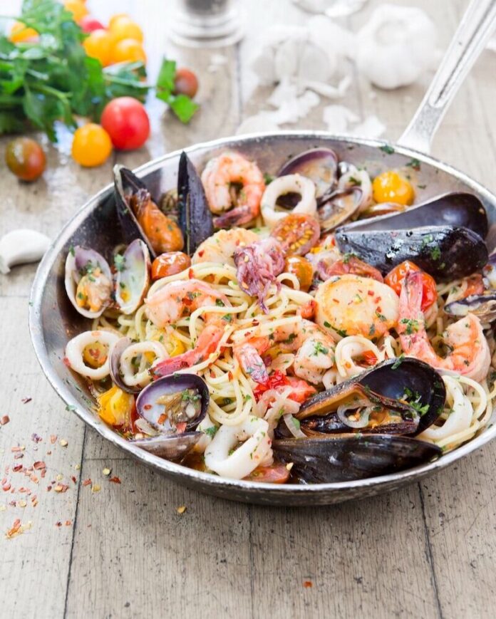 A seafood spaghetti sits in a shallow bowl with shrimp and mussels, all sitting on a wood picnic table.