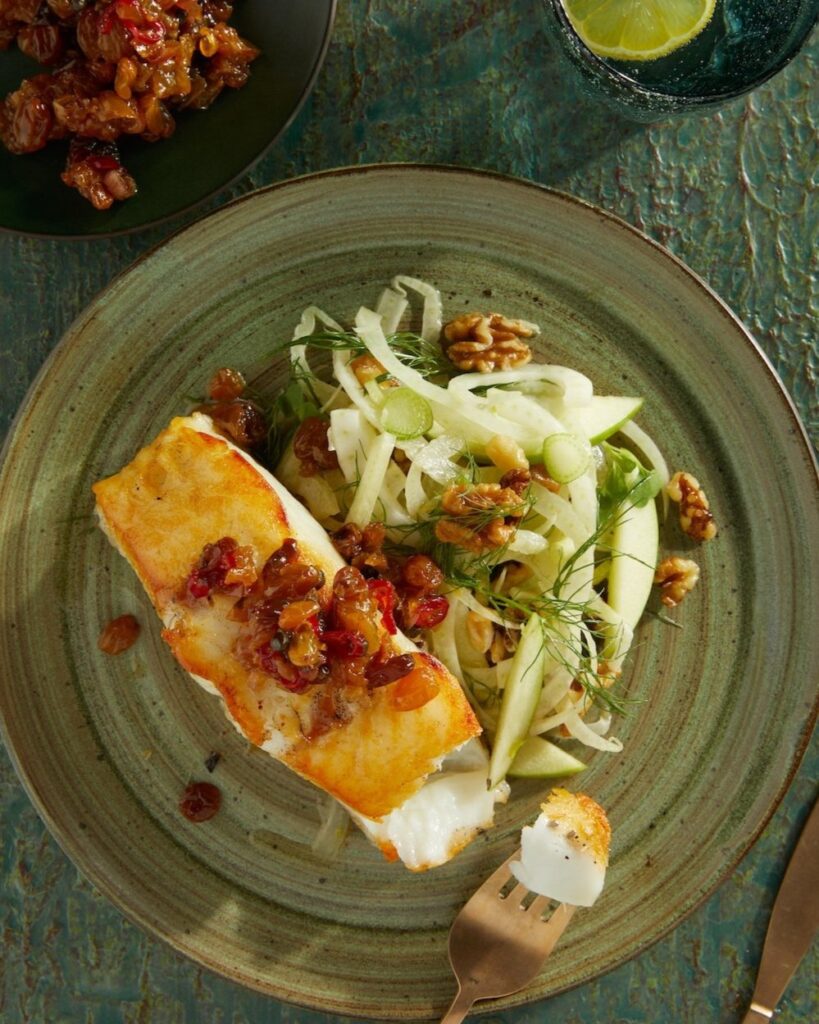 A piece of seared sea bass sits on a green plate with a raisin chutney and fennel salad beside it.