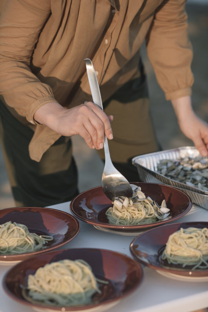 A person serves Spaghetti with Clams into four red bowls on a picnic table outside.