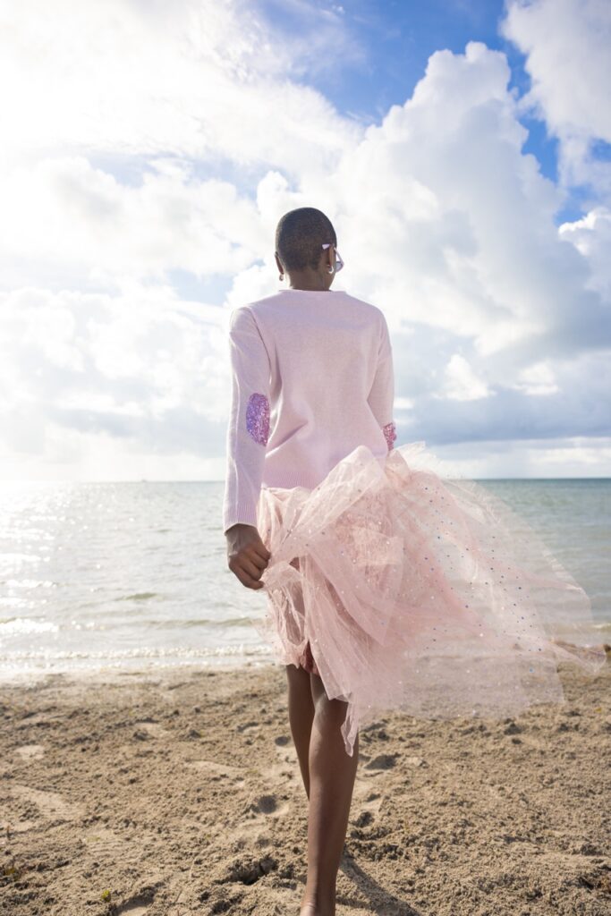 A woman in a pink long sleeve shirt and pink tutu walks on the beach towards the water.