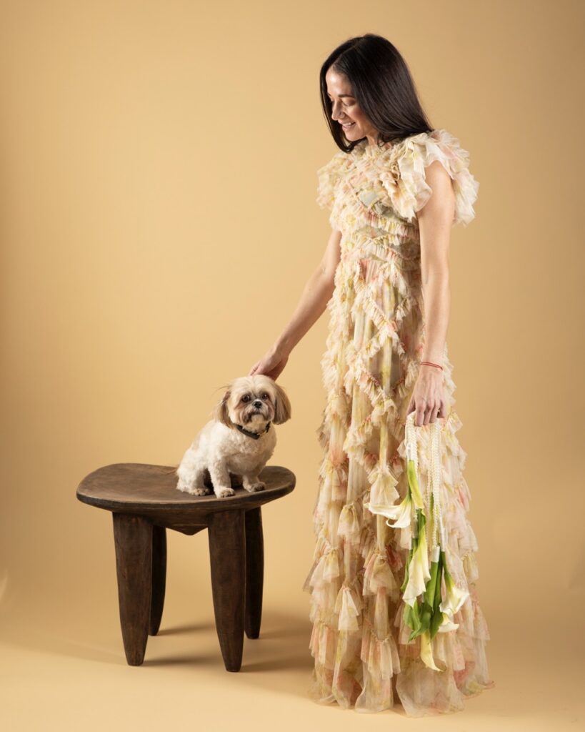 A woman wears a surrealism dress in cream while petting a dog on a stool.