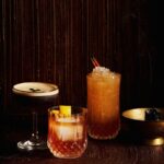 Three classic cocktail variations, an espresso martini, an old fashioned, and a mule sit on a dark table.