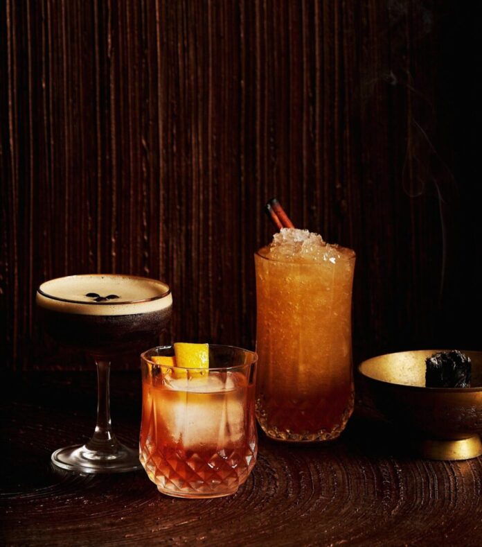 Three classic cocktail variations, an espresso martini, an old fashioned, and a mule sit on a dark table.