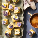 Small yellow triple citrus shortbread squares link a parchment paper baking sheet with dollops of whipped cream and purple edible flowers on top.