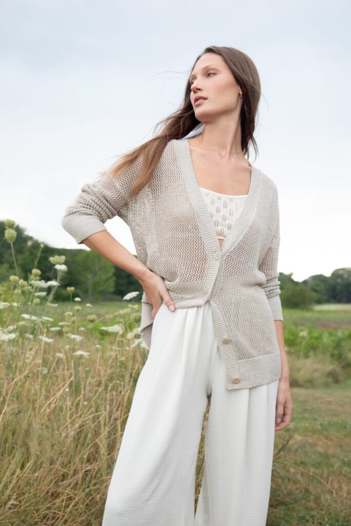 A woman stands in a field with a neutral cardigan and white underclothing on.