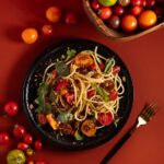 A bowl of BLT pasta with golden spaghetti, bacon pieces, red and yellow halved cherry tomatoes, and fresh green basil leaves.